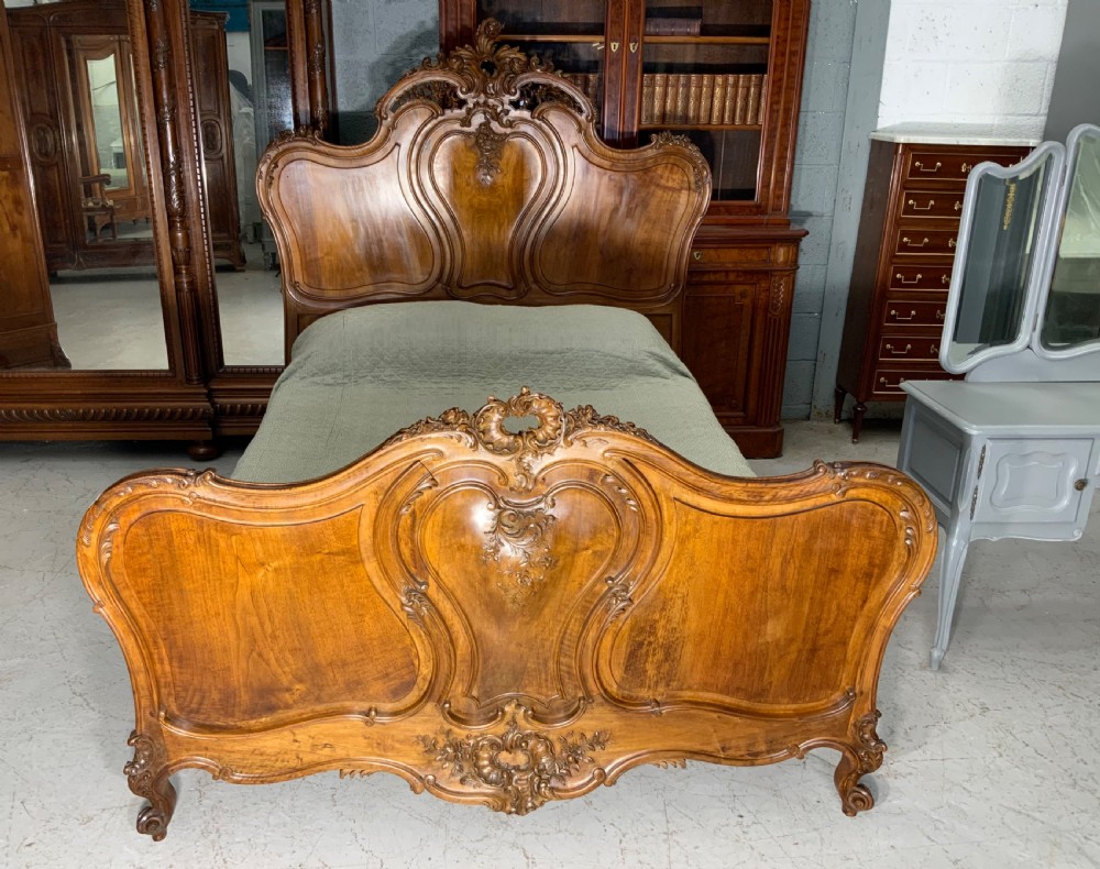 An antique carved walnut bed frame featuring an ornate arched headboard and footboard with carved scrollwork and floral cresting.
