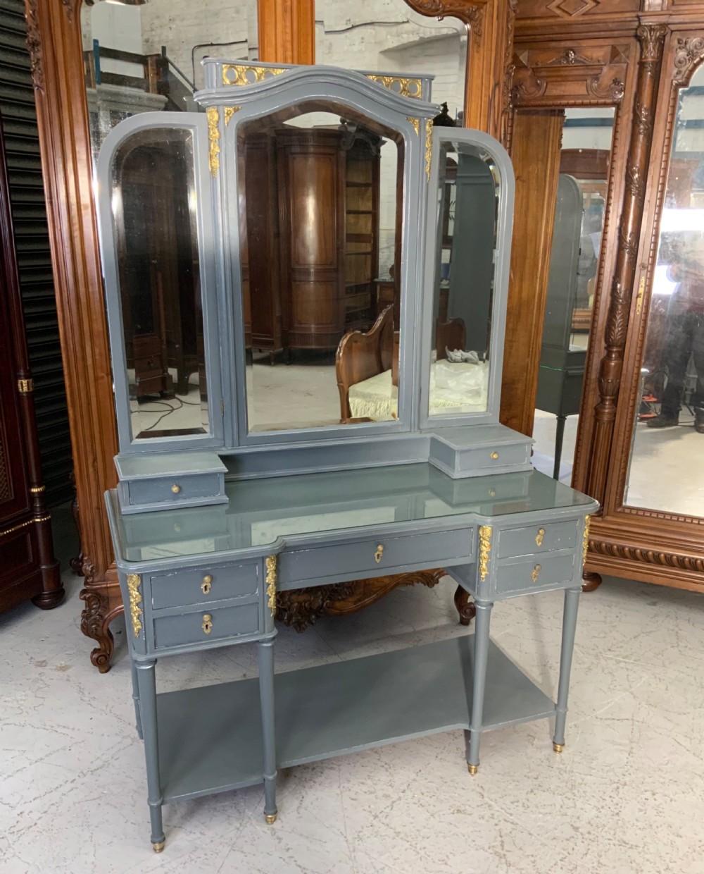 A plummet grey painted French dressing table featuring a central tri-fold mirror, brass embellishments, a glass top, multiple drawers, and turned legs.