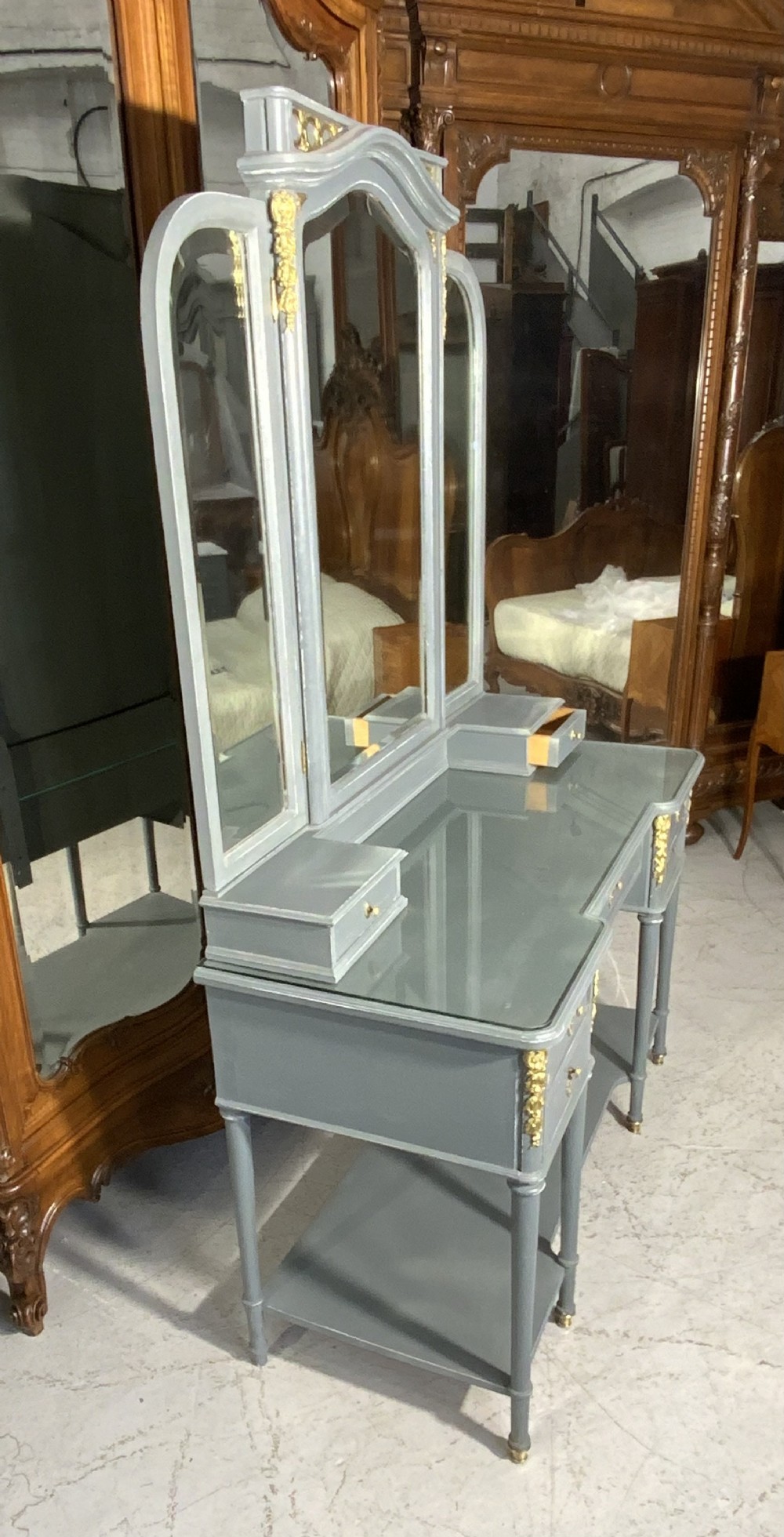 A plummet grey painted French dressing table featuring a central tri-fold mirror, brass embellishments, a glass top, multiple drawers, and turned legs. View IV