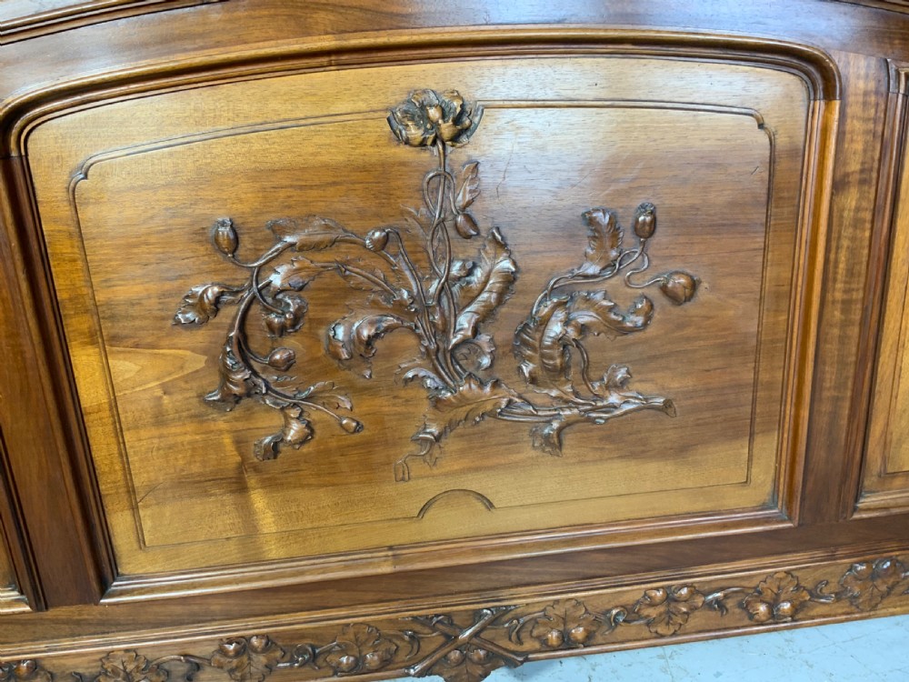 Carved walnut French bed frame with an arched headboard, ornate floral footboard carving, and a pair of matching wooden bedside cabinets. View X