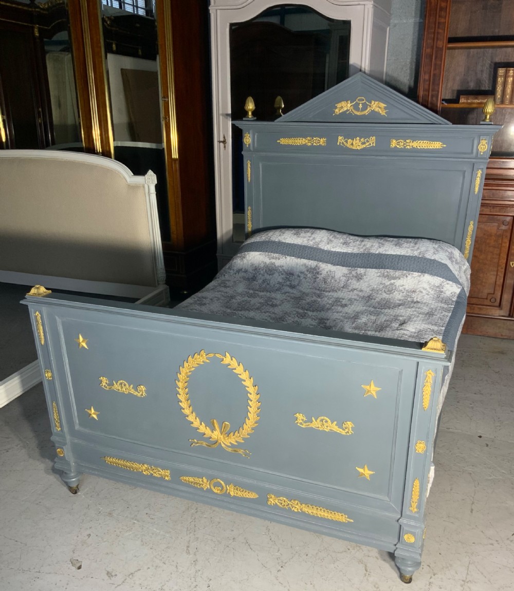 A grey painted French double bed frame decorated with gold brass mounts, featuring a central laurel wreath motif and a pediment headboard.