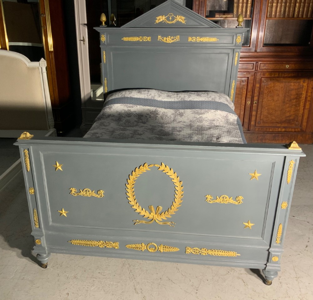 A grey painted French double bed frame decorated with gold brass mounts, featuring a central laurel wreath motif and a pediment headboard. View V