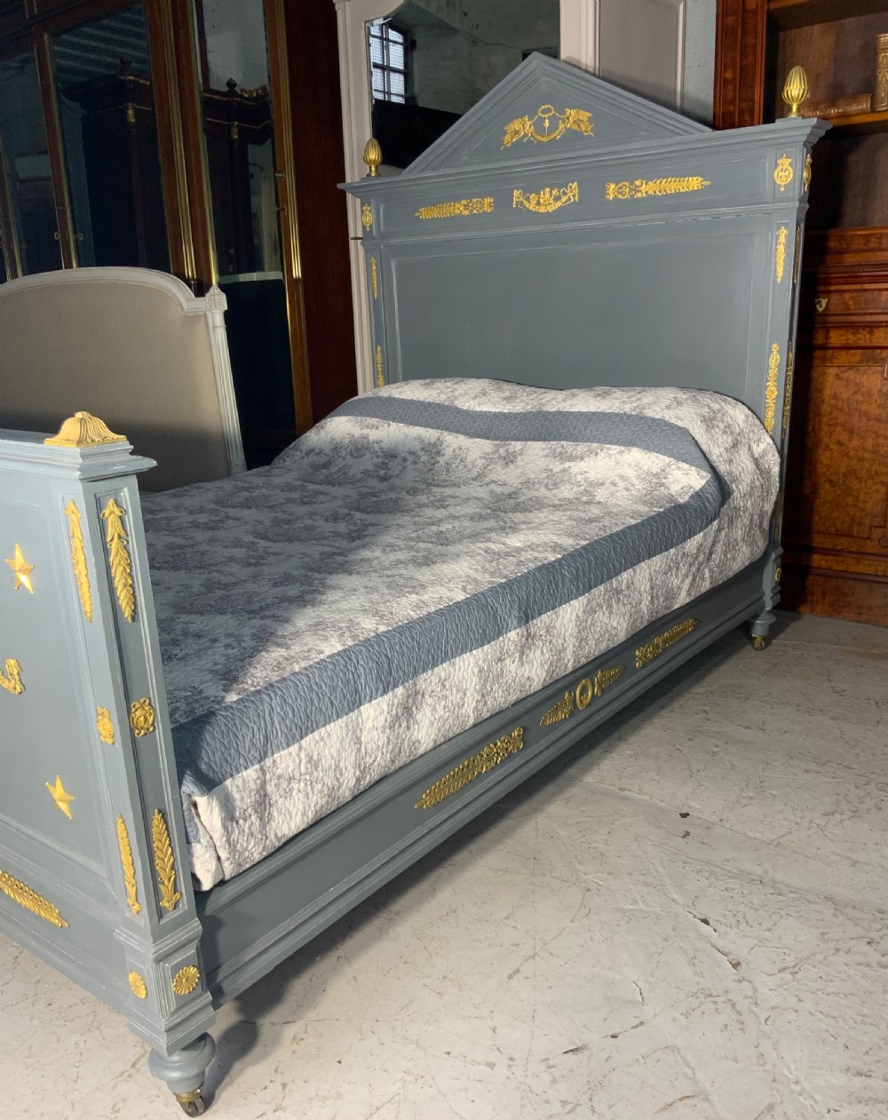 A grey painted French double bed frame decorated with gold brass mounts, featuring a central laurel wreath motif and a pediment headboard. View X