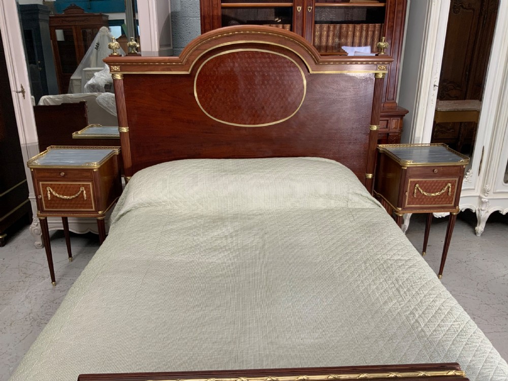 French Neoclassical mahogany bed frame featuring an oval marquetry panel, polished brass mounts, fluted columns, and two matching marble-topped bedside cabinets.