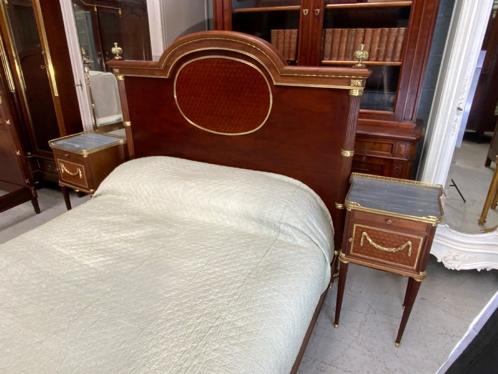 French Neoclassical mahogany bed frame featuring an oval marquetry panel, polished brass mounts, fluted columns, and two matching marble-topped bedside cabinets. View XV
