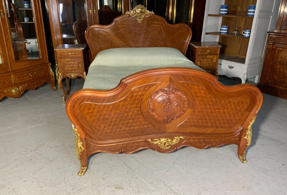 A French Louis XV style king-size bedstead featuring geometric parquetry inlay, carved central motif, and gilt-bronze mounts, flanked by two matching bedside cabinets with marble tops.