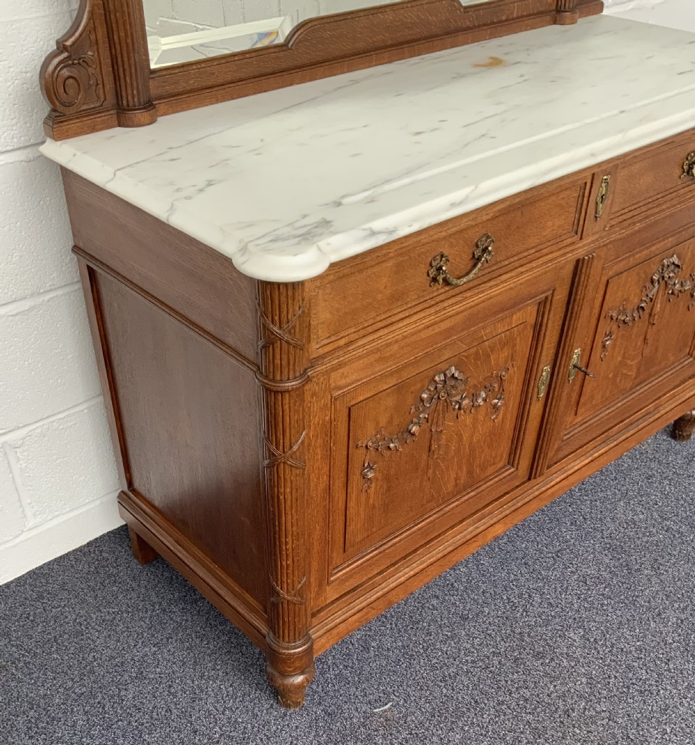 A carved oak washstand featuring a white marble countertop, a large arched mirror framed in oak with top crest carving, two drawers, and a double-door cabinet below. View IV
