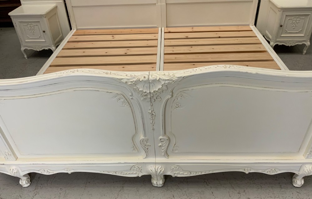 A cream painted French Rococo style bedstead featuring detailed foliage carving, serpentine headboard, matching footboard, and two bedside cabinets. View III
