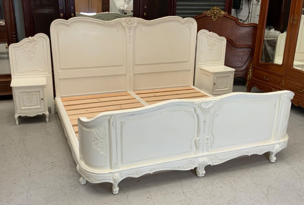 A cream painted French Rococo style bedstead featuring detailed foliage carving, serpentine headboard, matching footboard, and two bedside cabinets. View XIX