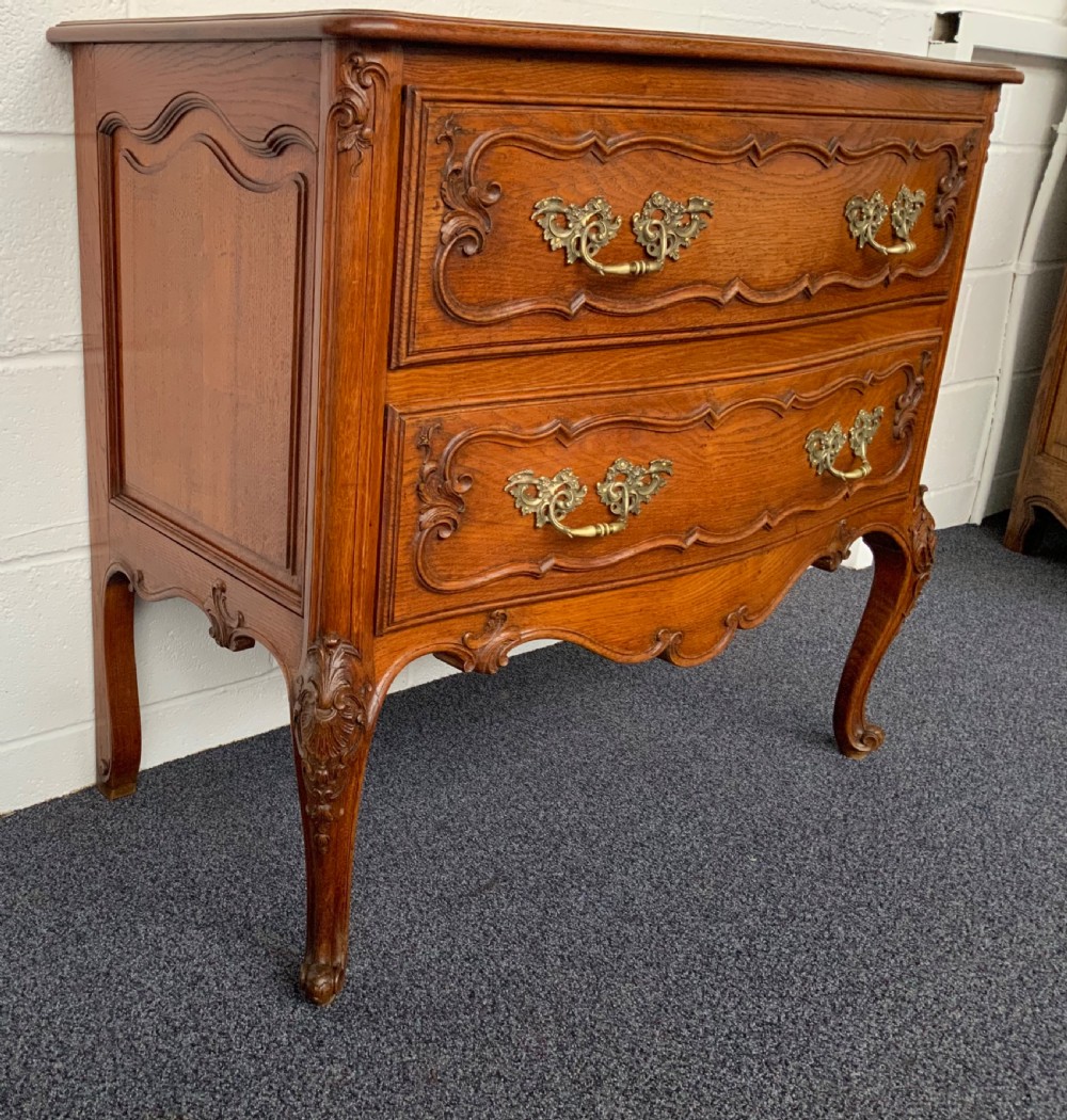 A carved oak French bow-front chest of drawers featuring two drawers with ornate brass handles, a shaped apron, and cabriole legs. View III