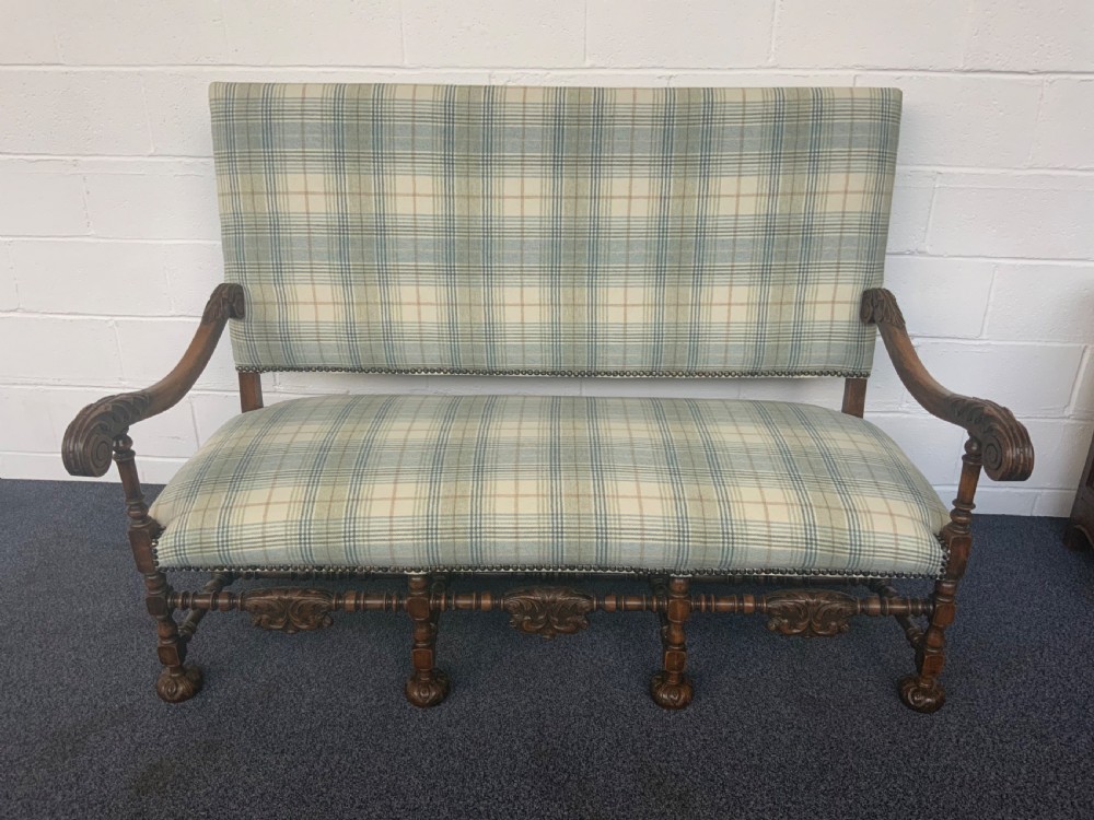 An upholstered French carved walnut settee featuring a green and cream check fabric, carved scroll arms, turned stretchers, and brass studding.