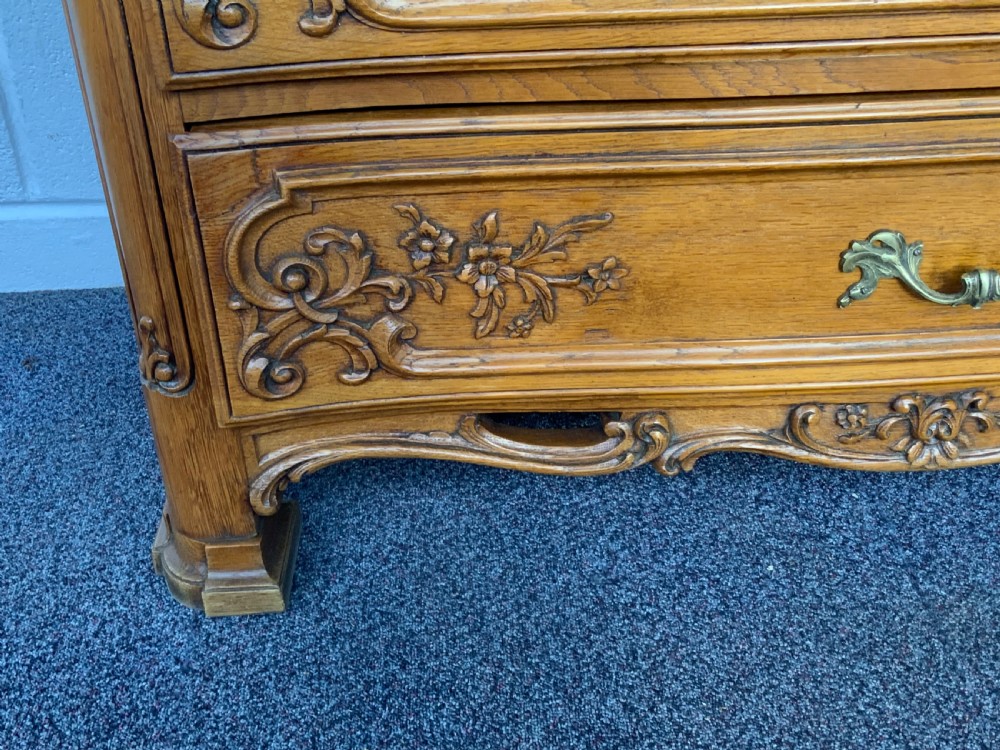 A French Provincial style warm oak chest of drawers featuring three wide drawers with floral relief carvings and brass scroll handles above a scalloped apron. View III