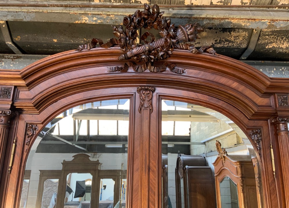 A tall 19th-century French rosewood armoire with two full-length mirrored doors, an arched pediment topped with a foliage crest carving, and fluted corner columns. View III