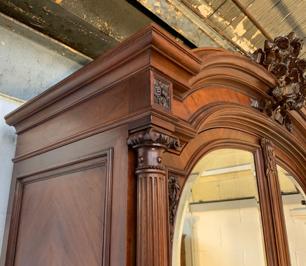 A tall 19th-century French rosewood armoire with two full-length mirrored doors, an arched pediment topped with a foliage crest carving, and fluted corner columns. View VI
