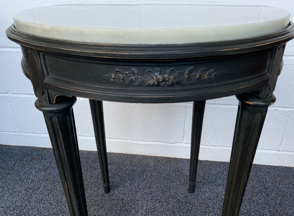 A circular French centre table featuring a light grey marble top set upon a dark painted carved frame with tapered fluted legs. View III