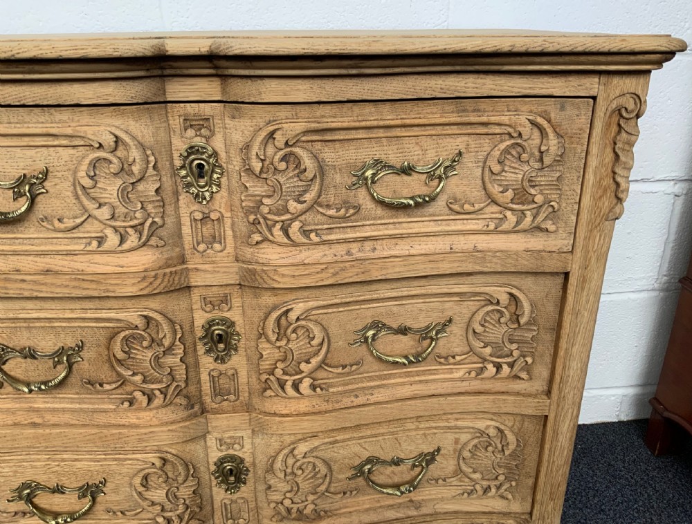 A light bleached oak French commode featuring three carved drawer fronts, brass handles, and short cabriole legs. View V