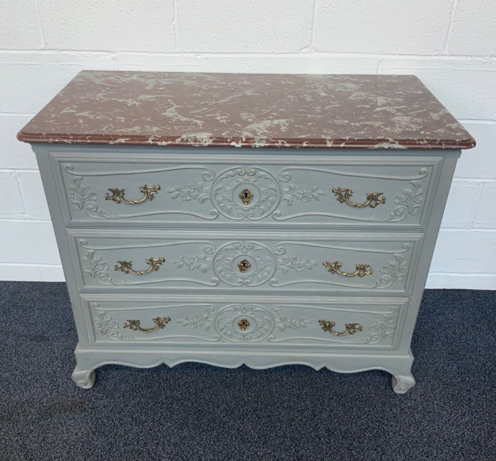 A grey painted French chest of drawers featuring three carved drawers, brass handles, cabriole legs, and a red variegated marble top.