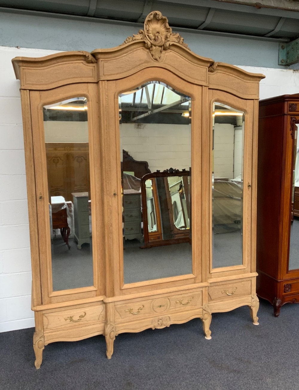 A large triple-door bleached oak French armoire featuring arched mirror panels, carved rococo cresting, bottom drawers, and cabriole feet.