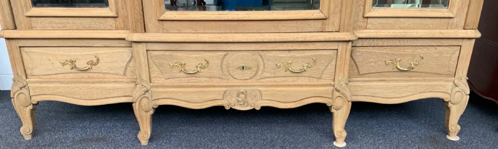 A large triple-door bleached oak French armoire featuring arched mirror panels, carved rococo cresting, bottom drawers, and cabriole feet. View III