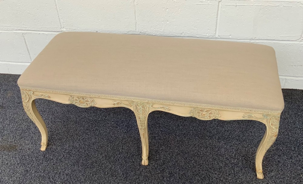 A long rectangular French-style dressing stool featuring a distressed cream painted wooden frame, carved cabriole legs, and neutral beige linen upholstery.