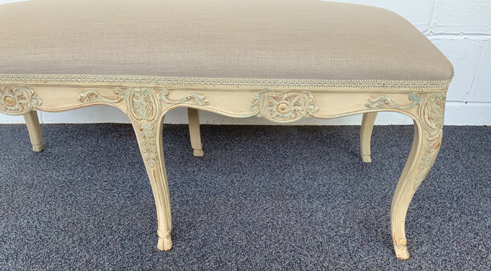 A long rectangular French-style dressing stool featuring a distressed cream painted wooden frame, carved cabriole legs, and neutral beige linen upholstery. View II