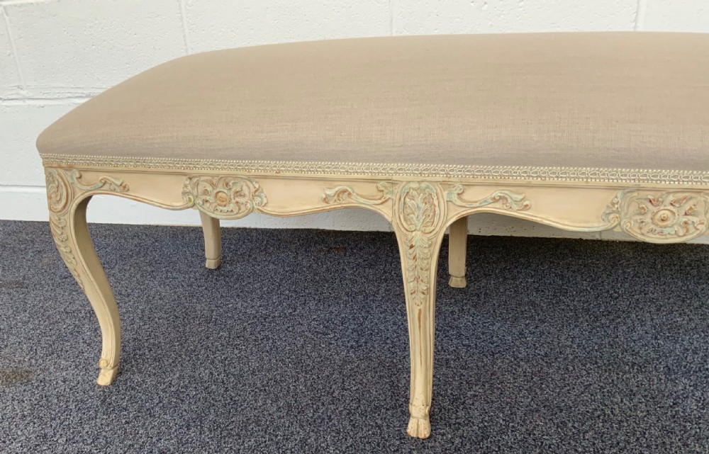 A long rectangular French-style dressing stool featuring a distressed cream painted wooden frame, carved cabriole legs, and neutral beige linen upholstery. View III