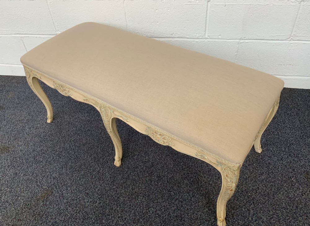A long rectangular French-style dressing stool featuring a distressed cream painted wooden frame, carved cabriole legs, and neutral beige linen upholstery. View VI