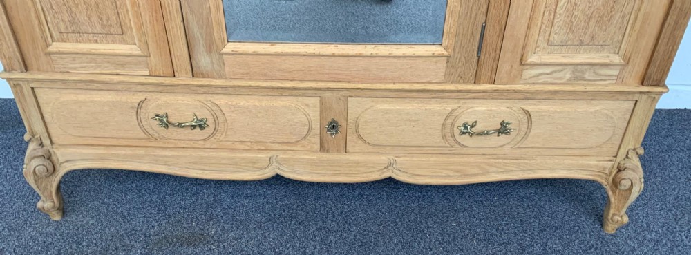 A large bleached oak triple-door armoire featuring glass mirror panels, a carved top crest pediment, base drawers, and carved cabriole feet. View II