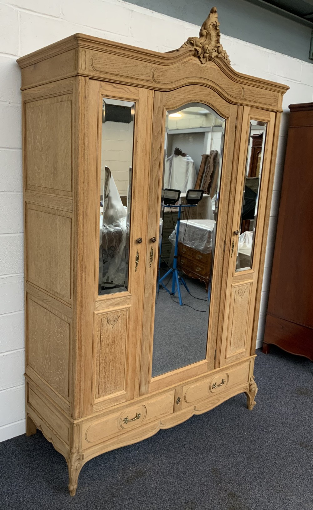 A large bleached oak triple-door armoire featuring glass mirror panels, a carved top crest pediment, base drawers, and carved cabriole feet. View VI