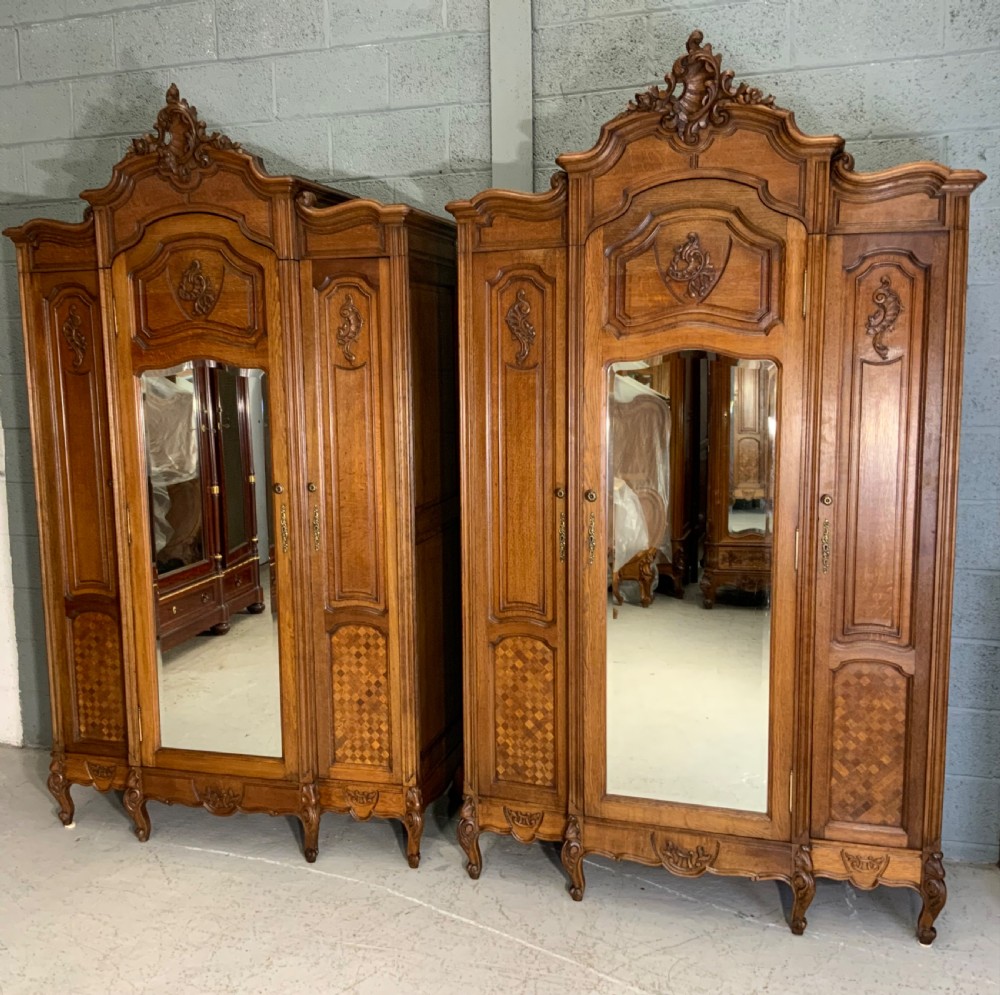 A pair of carved oak French provincial armoires featuring arched mirrored doors, parquetry lower panels, and elaborate carved crests.