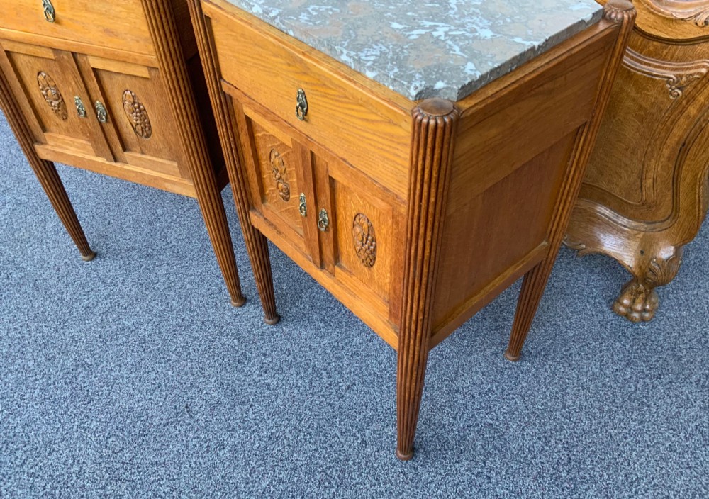 A pair of French Art Deco oak bedside cabinets featuring inset grey marble tops, single upper drawers, carved door panels, and tapered fluted legs. View IV