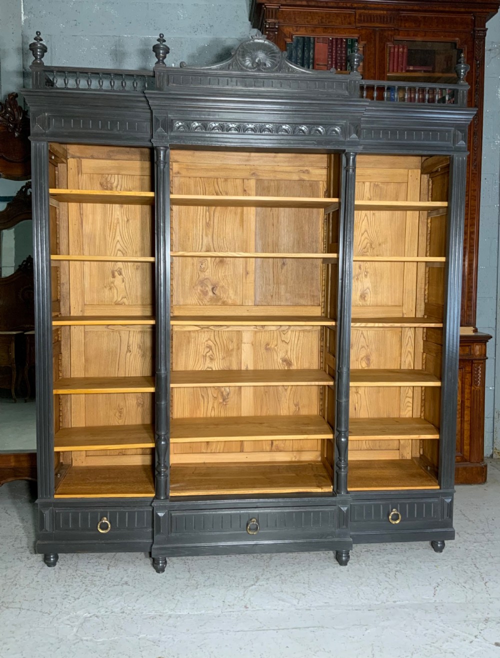 A large antique French open bookcase featuring a dark painted frame with carved shell cresting and finials, contrasting natural pine shelving, and three bottom drawers with brass handles.