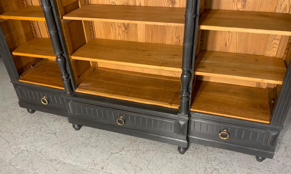 A large antique French open bookcase featuring a dark painted frame with carved shell cresting and finials, contrasting natural pine shelving, and three bottom drawers with brass handles. View II