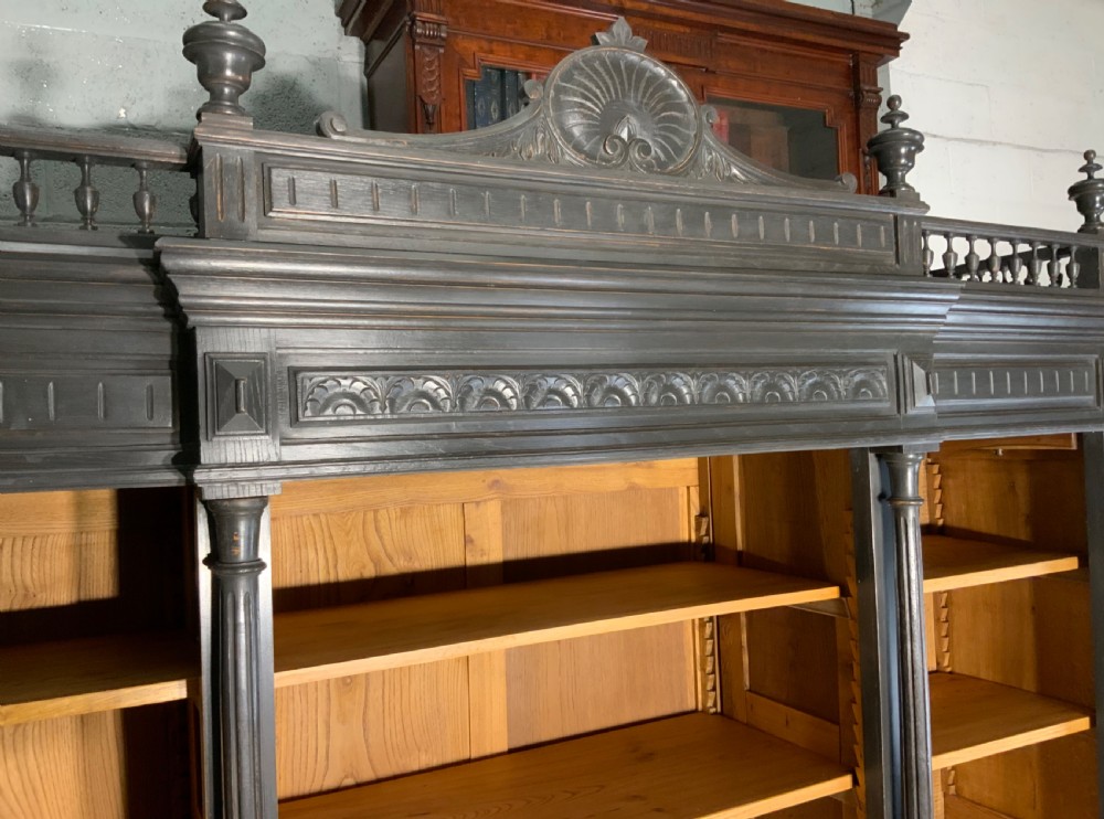 A large antique French open bookcase featuring a dark painted frame with carved shell cresting and finials, contrasting natural pine shelving, and three bottom drawers with brass handles. View VII