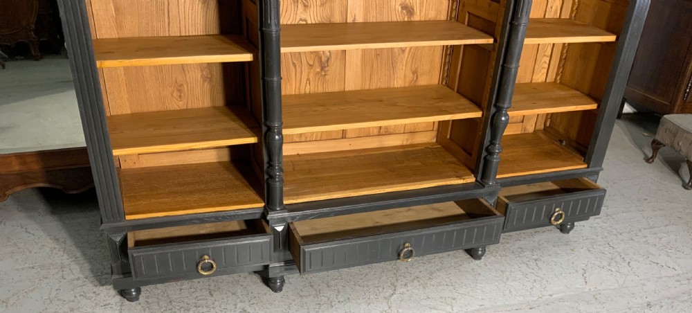 A large antique French open bookcase featuring a dark painted frame with carved shell cresting and finials, contrasting natural pine shelving, and three bottom drawers with brass handles. View X