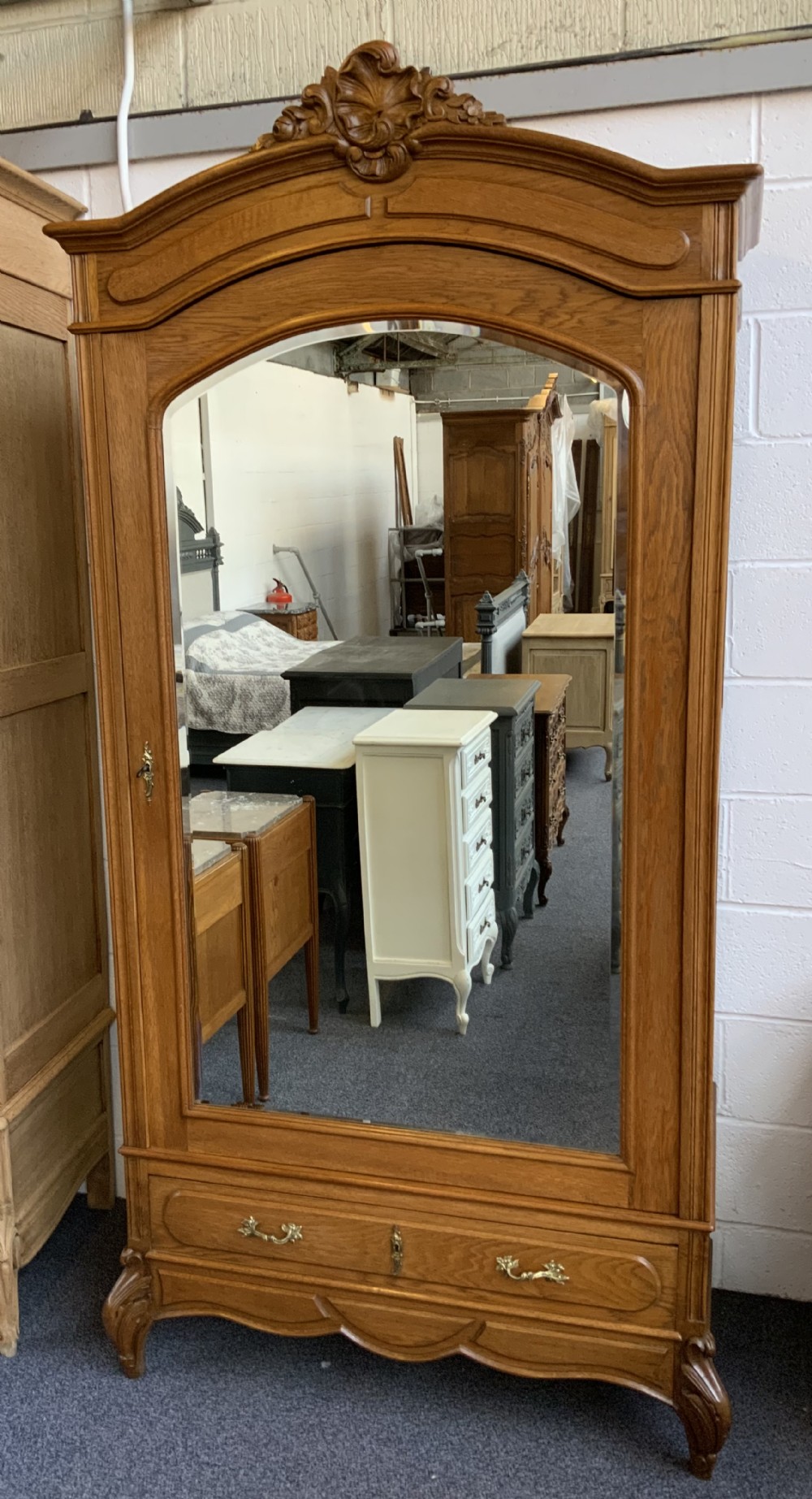 A tall French oak single-door armoire featuring an arched mirrored door, a carved cresting pediment, a bottom drawer, and cabriole feet.