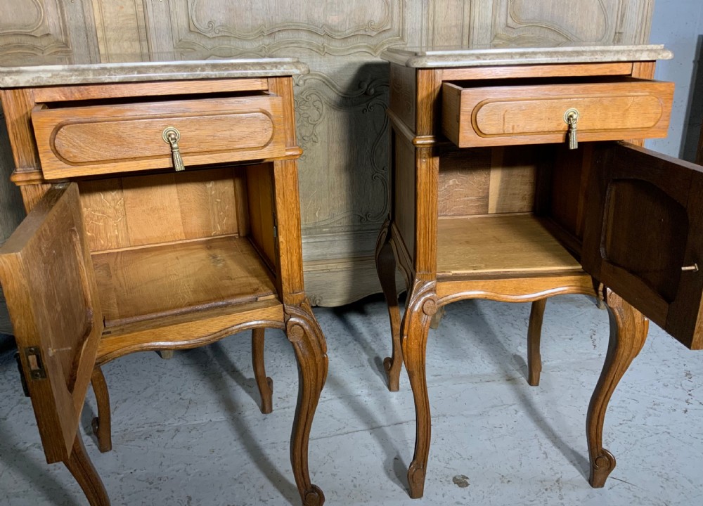 A matching pair of French oak bedside cabinets featuring inset beige marble tops, single frieze drawers, carved cupboard doors, and slender cabriole legs. View II