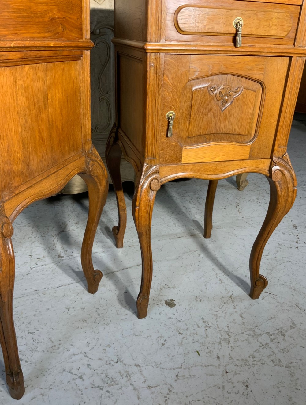 A matching pair of French oak bedside cabinets featuring inset beige marble tops, single frieze drawers, carved cupboard doors, and slender cabriole legs. View VI