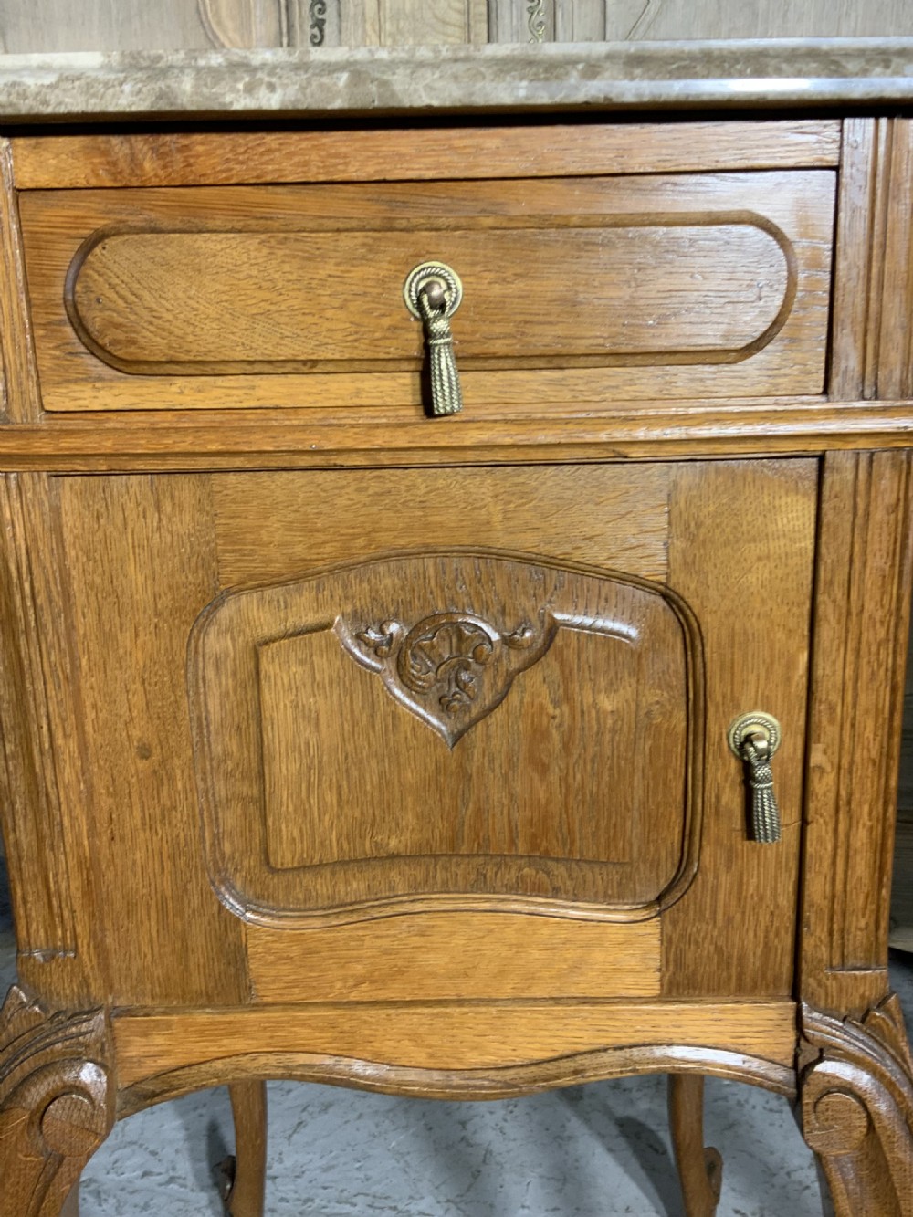 A matching pair of French oak bedside cabinets featuring inset beige marble tops, single frieze drawers, carved cupboard doors, and slender cabriole legs. View VII