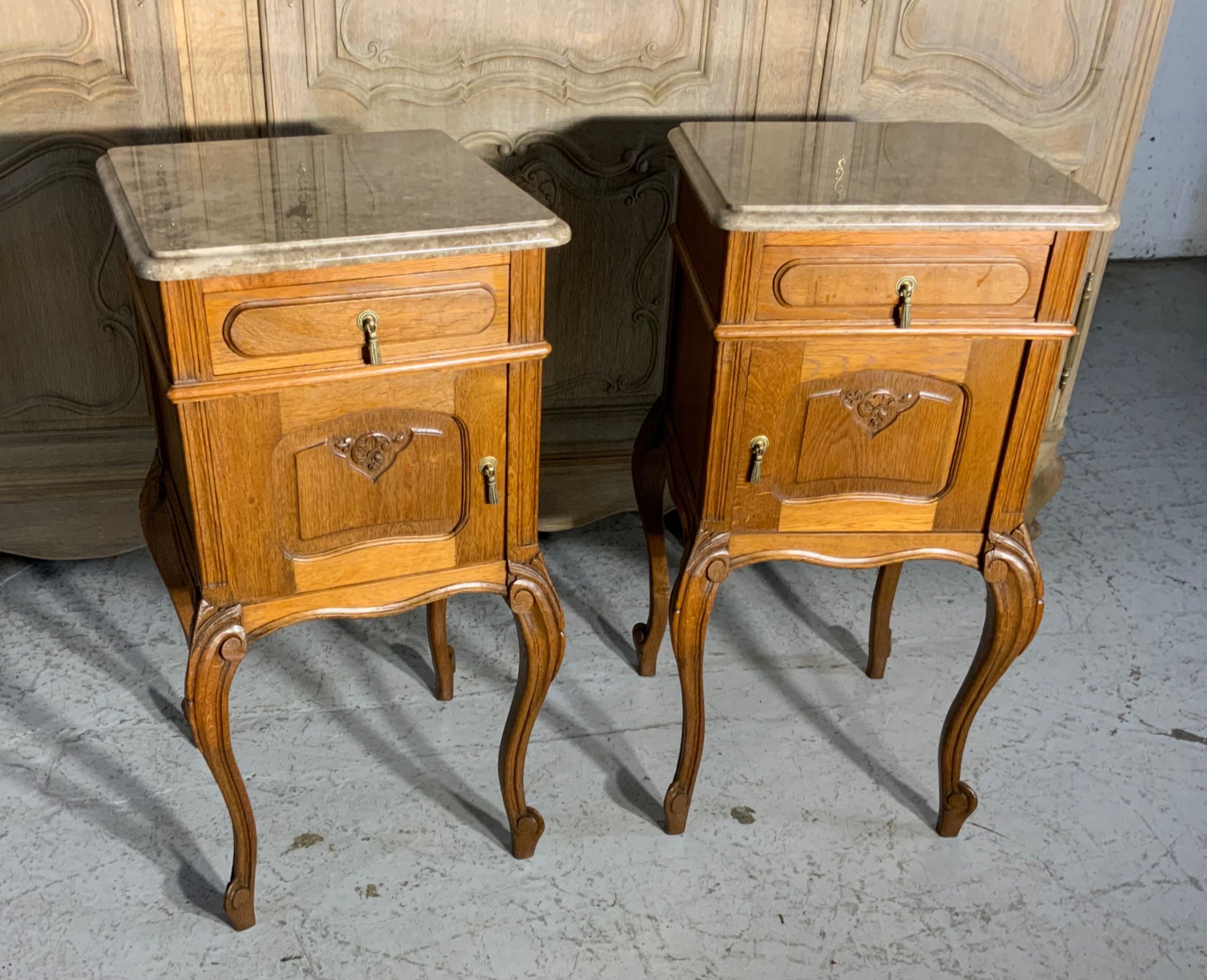 A matching pair of French oak bedside cabinets featuring inset beige marble tops, single frieze drawers, carved cupboard doors, and slender cabriole legs.