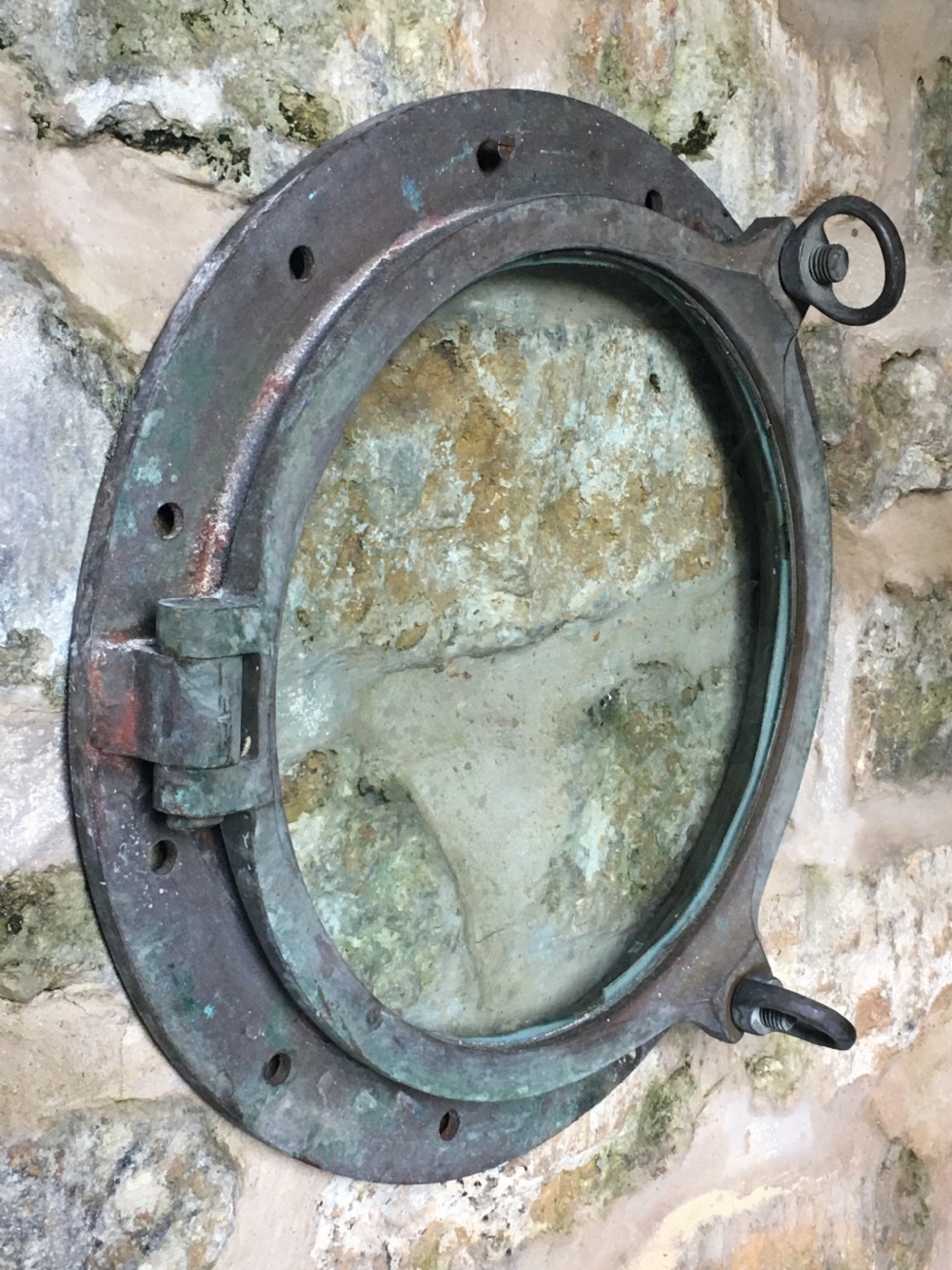 A heavy antique bronze circular ship porthole featuring visible hinge mounts, dog-bolt fasteners, a clear glass pane, and natural green patina.