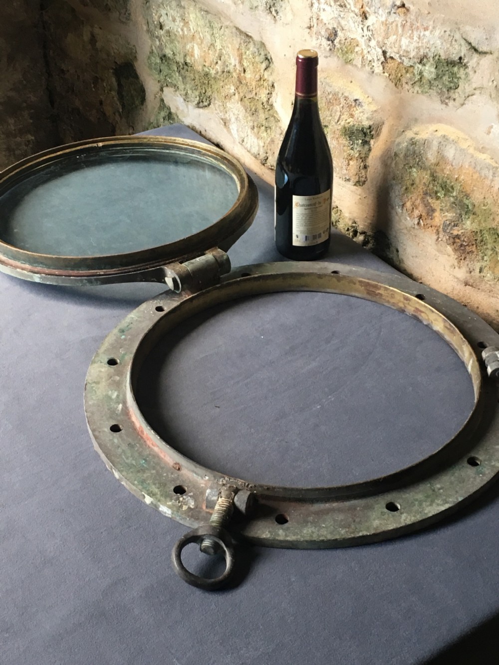 A heavy antique bronze circular ship porthole featuring visible hinge mounts, dog-bolt fasteners, a clear glass pane, and natural green patina. View II