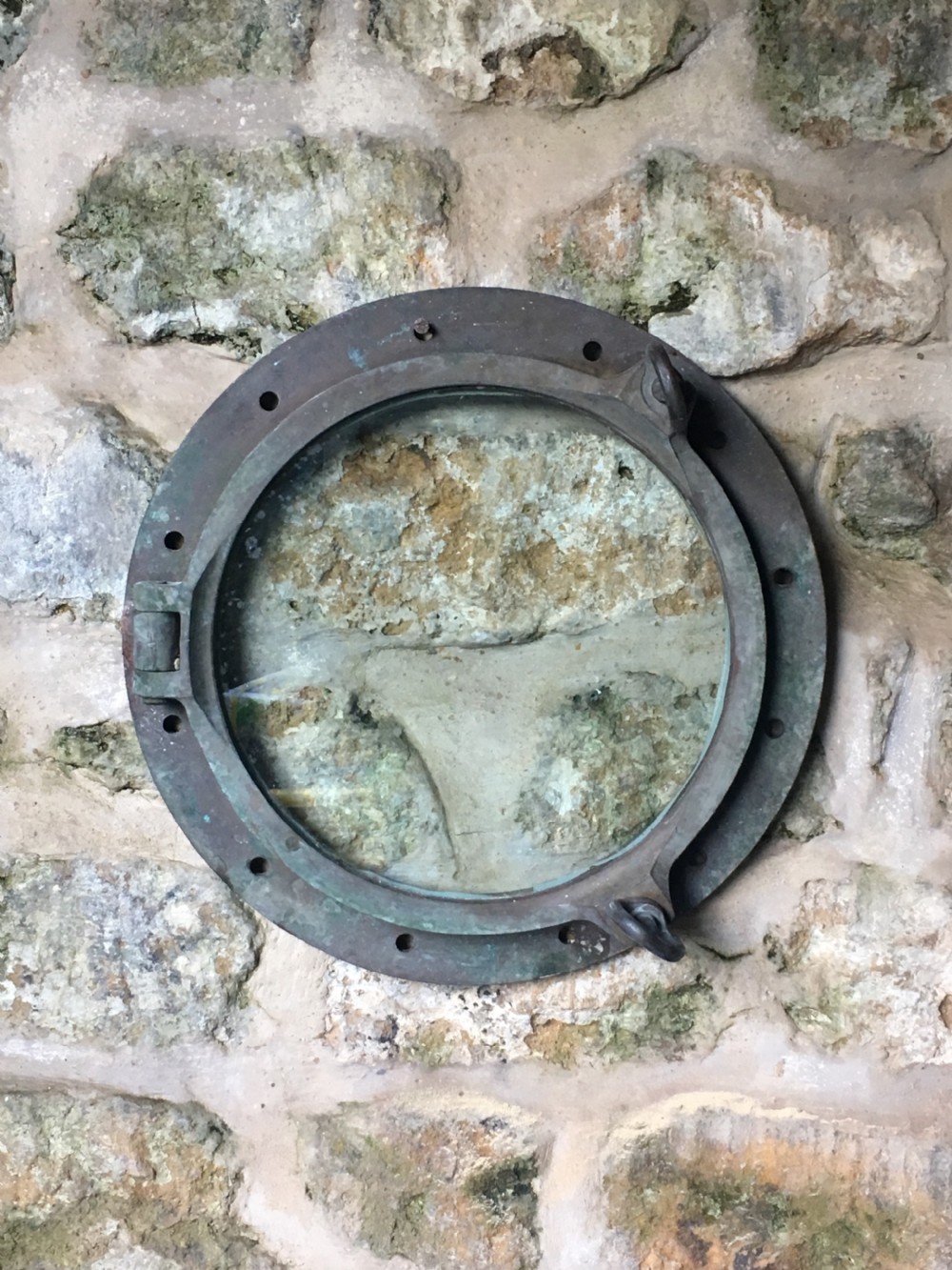 A heavy antique bronze circular ship porthole featuring visible hinge mounts, dog-bolt fasteners, a clear glass pane, and natural green patina. View III