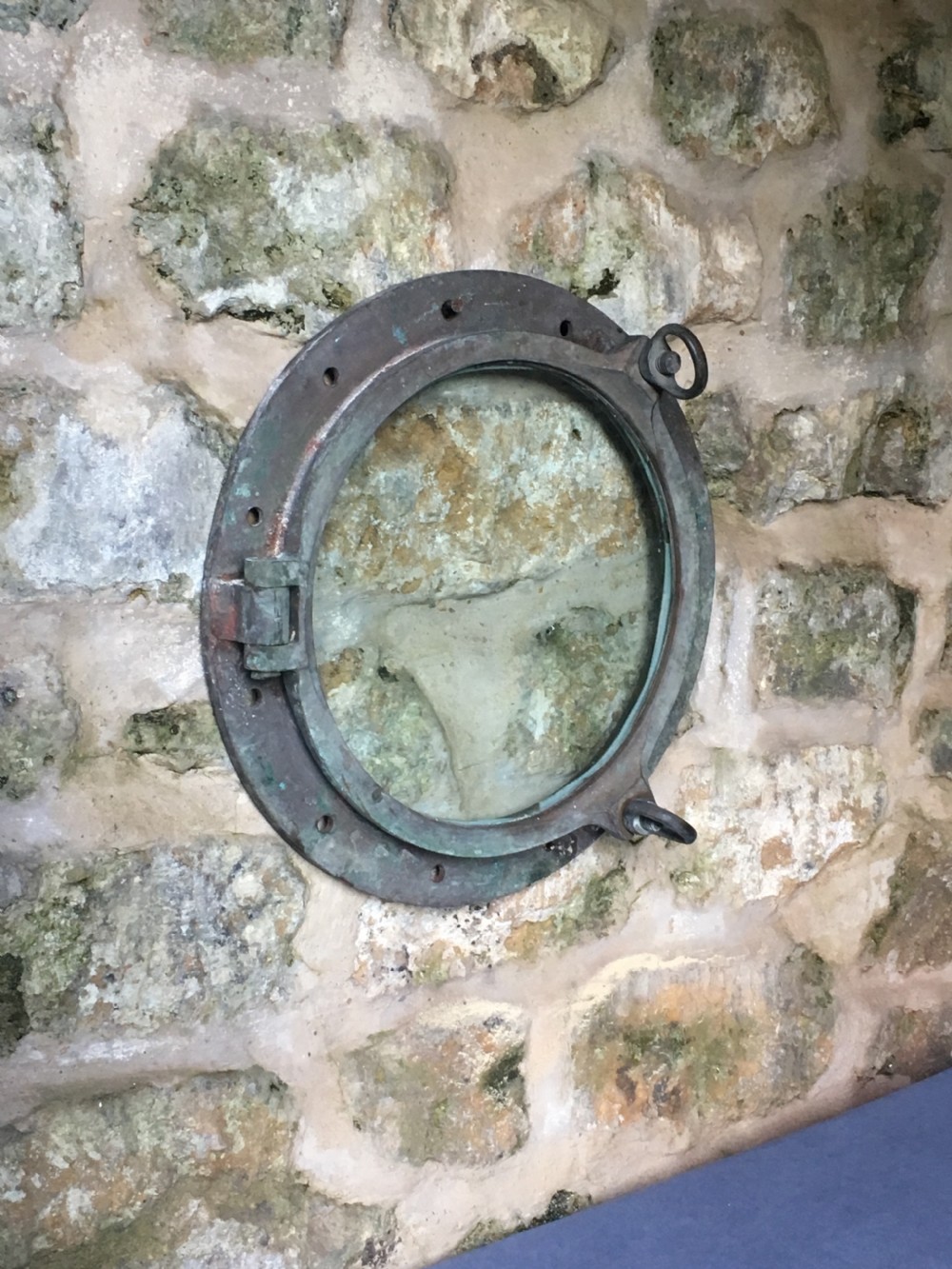 A heavy antique bronze circular ship porthole featuring visible hinge mounts, dog-bolt fasteners, a clear glass pane, and natural green patina. View V
