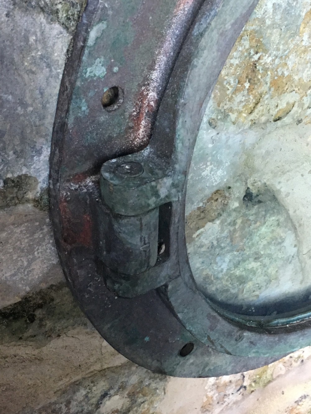 A heavy antique bronze circular ship porthole featuring visible hinge mounts, dog-bolt fasteners, a clear glass pane, and natural green patina. View VI
