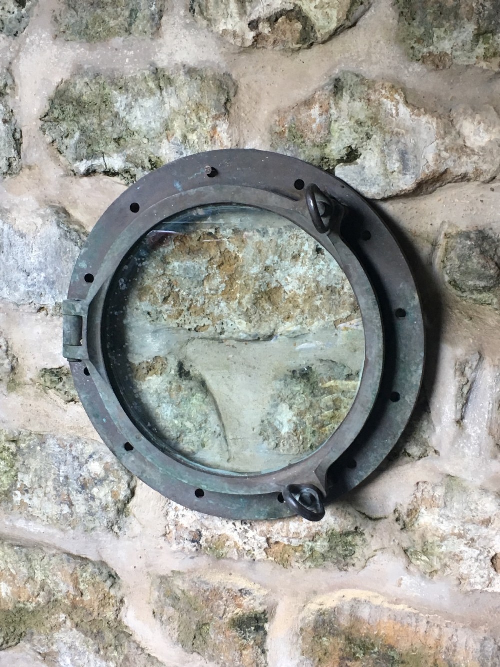 A heavy antique bronze circular ship porthole featuring visible hinge mounts, dog-bolt fasteners, a clear glass pane, and natural green patina. View VII