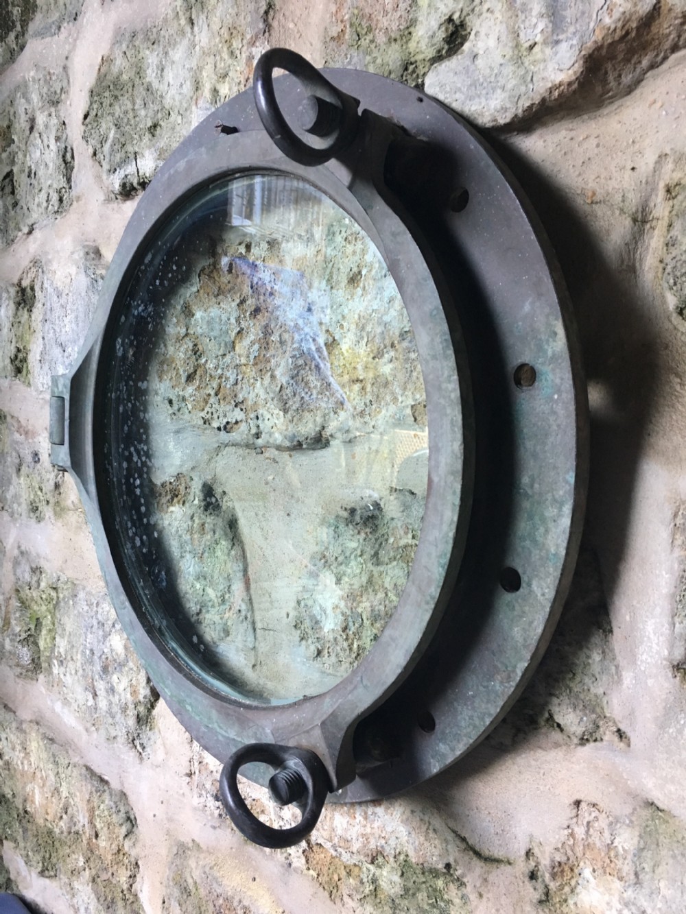 A heavy antique bronze circular ship porthole featuring visible hinge mounts, dog-bolt fasteners, a clear glass pane, and natural green patina. View IX