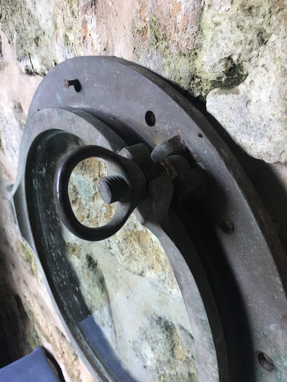 A heavy antique bronze circular ship porthole featuring visible hinge mounts, dog-bolt fasteners, a clear glass pane, and natural green patina. View X
