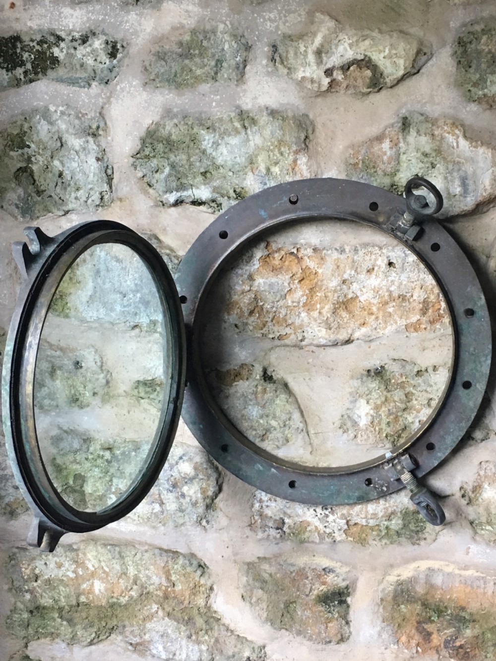 A heavy antique bronze circular ship porthole featuring visible hinge mounts, dog-bolt fasteners, a clear glass pane, and natural green patina. View XII