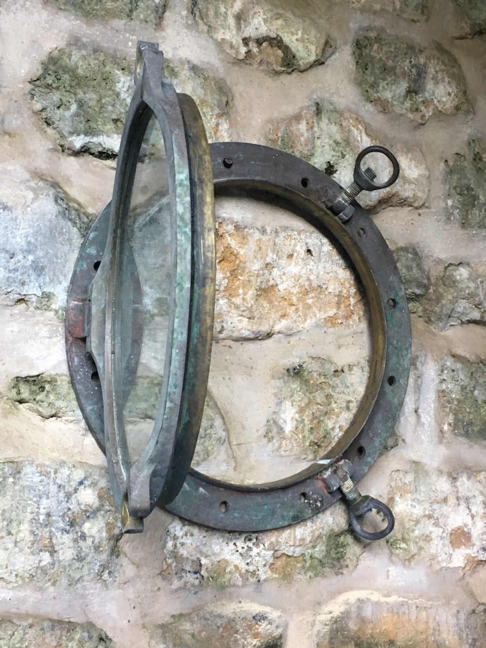 A heavy antique bronze circular ship porthole featuring visible hinge mounts, dog-bolt fasteners, a clear glass pane, and natural green patina. View XIII